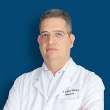 Médico de óculos e jaleco branco posando para retrato profissional sobre fundo azul sólido.